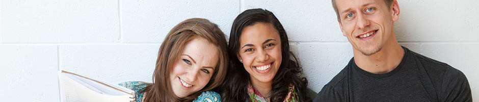 Two female students and a male student sitting against a white concrete wall smiling