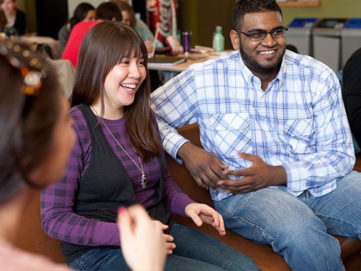 A young male and female student seated side-by-side, looking off-camera and laughing