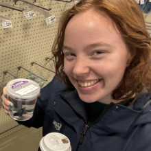 Alyssa holding drinks in front of hooks in a hardware store.