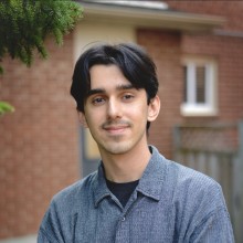 Davian standing in front of a red brick house and a hence. There is an evergreen tree off to the side above his head.
